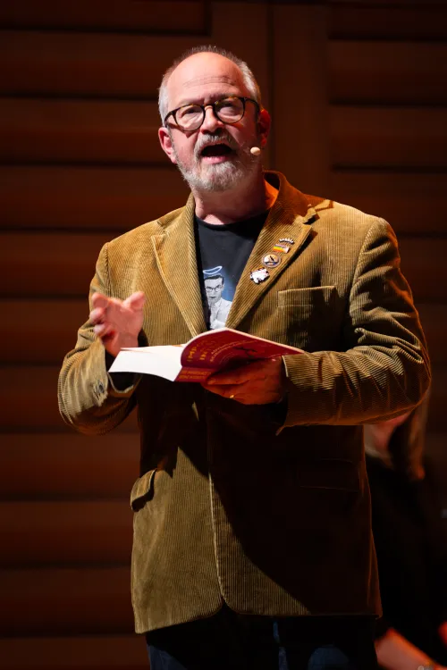 Robin Ince wearing brown corduroy jacket while reading a book on stage. 