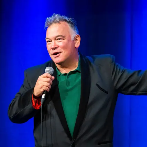 Stuart Lee holding a microphone while stood against a bright blue background. 